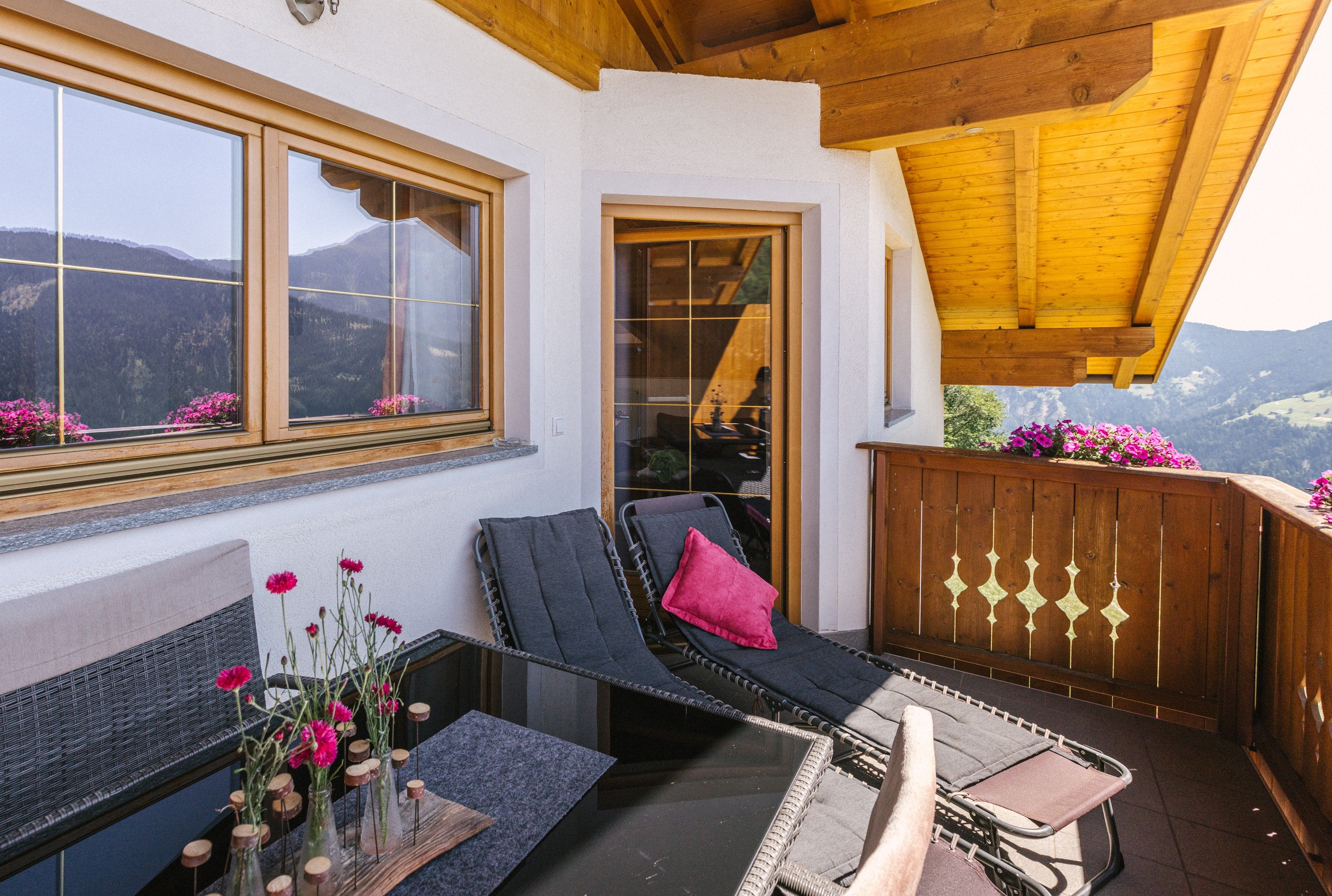 Un balcone con vista sulle montagne, una sedia in vimini con un cuscino, un tavolino con un vaso di fiori e un recinto in legno.