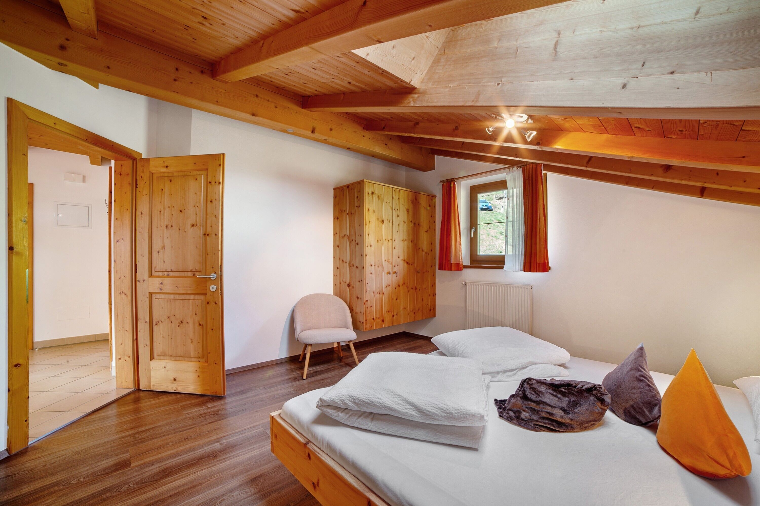 Techo de madera, una cama con almohadas, una silla y una ventana con cortinas.