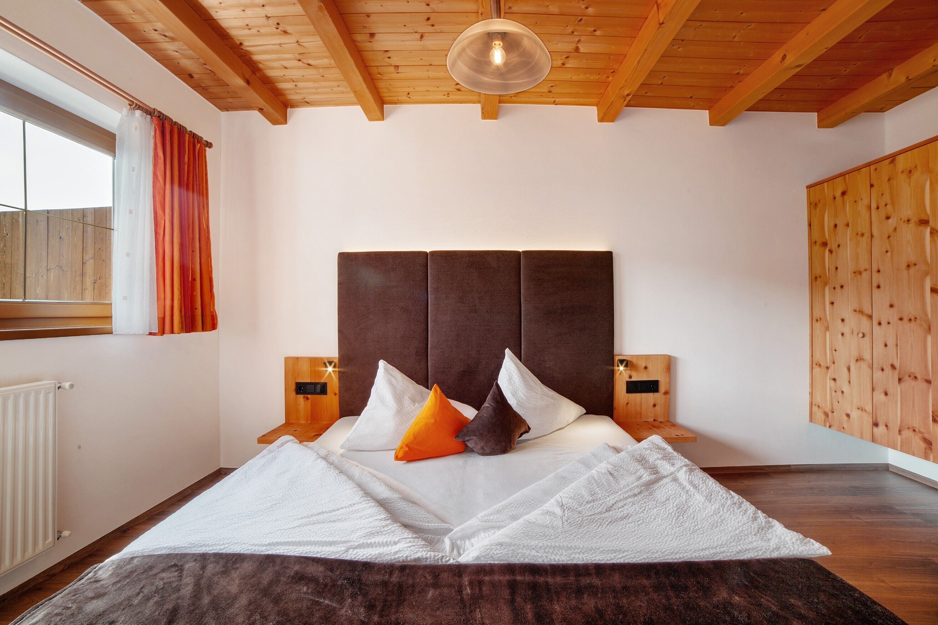 A bedroom with a wooden ceiling, a large bed with white and orange pillows, wooden nightstands, and a window with curtains.