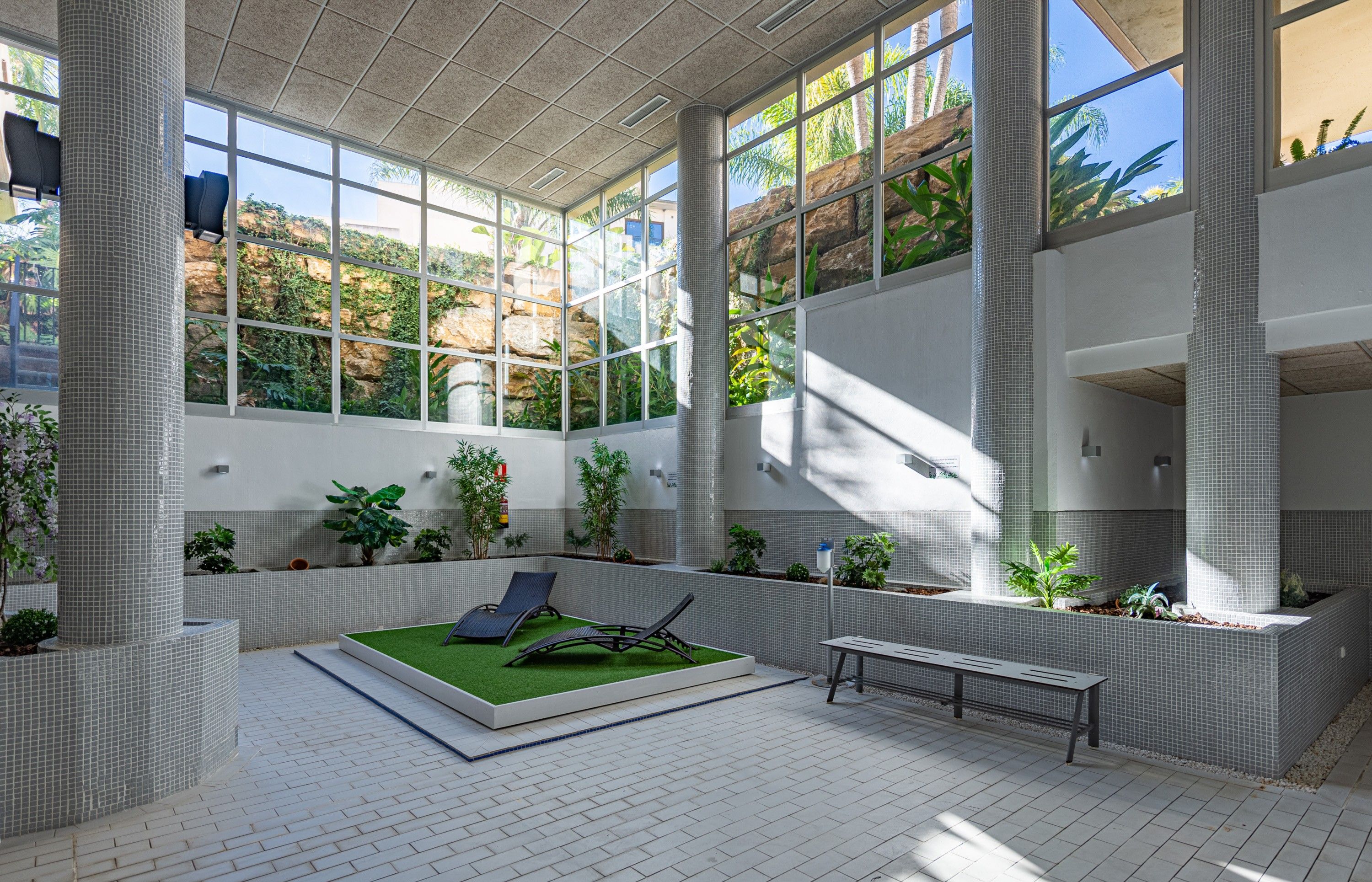 Un vaste atrium doté de grandes fenêtres, de plantes d’intérieur et d’un espace de détente central agrémenté d’une petite portion de gazon artificiel.