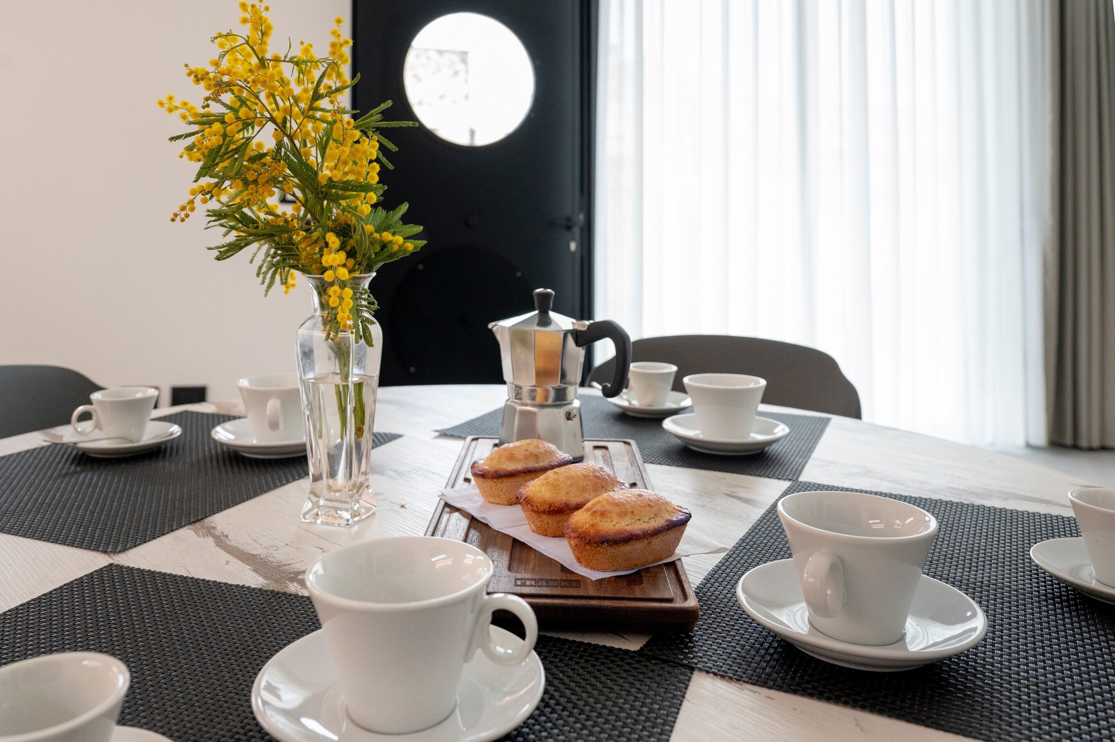 A dining table with white cups, saucers, a clear vase with yellow flowers, and a tray with muffins.