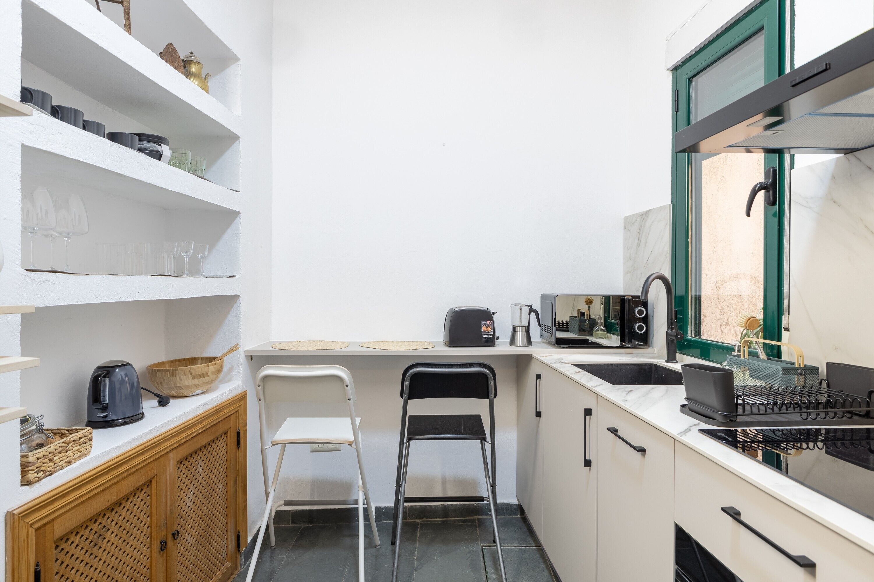 Una cocina compacta con armarios blancos, una encimera negra, un ventanal con marco verde y un fregadero.