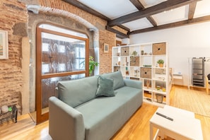 A room with a light grey sofa, a white side table, a white shelving unit with decorative items, a brick wall, and a wooden ceiling.