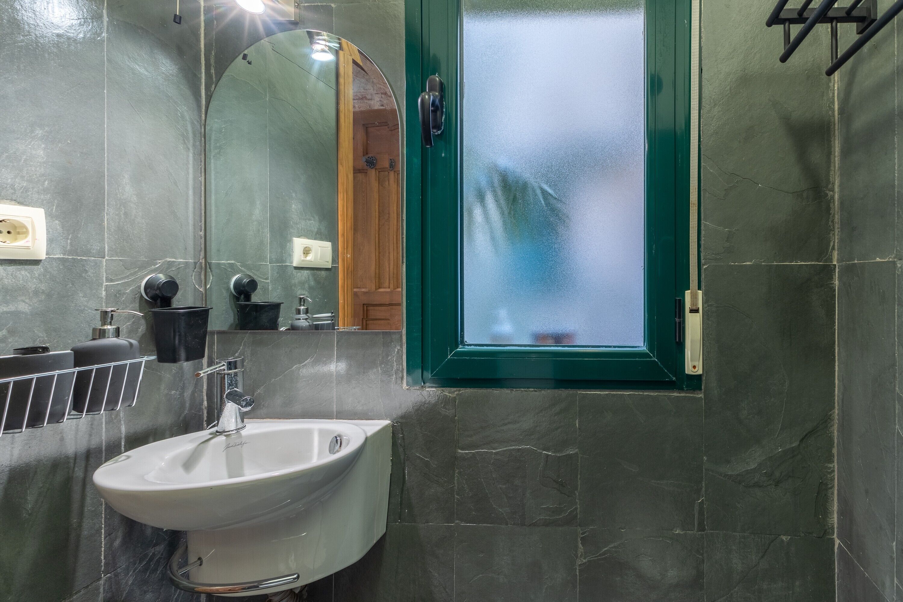 Un baño con un lavamanos blanco, un espejo, una ventana con marco verde y un perchero.