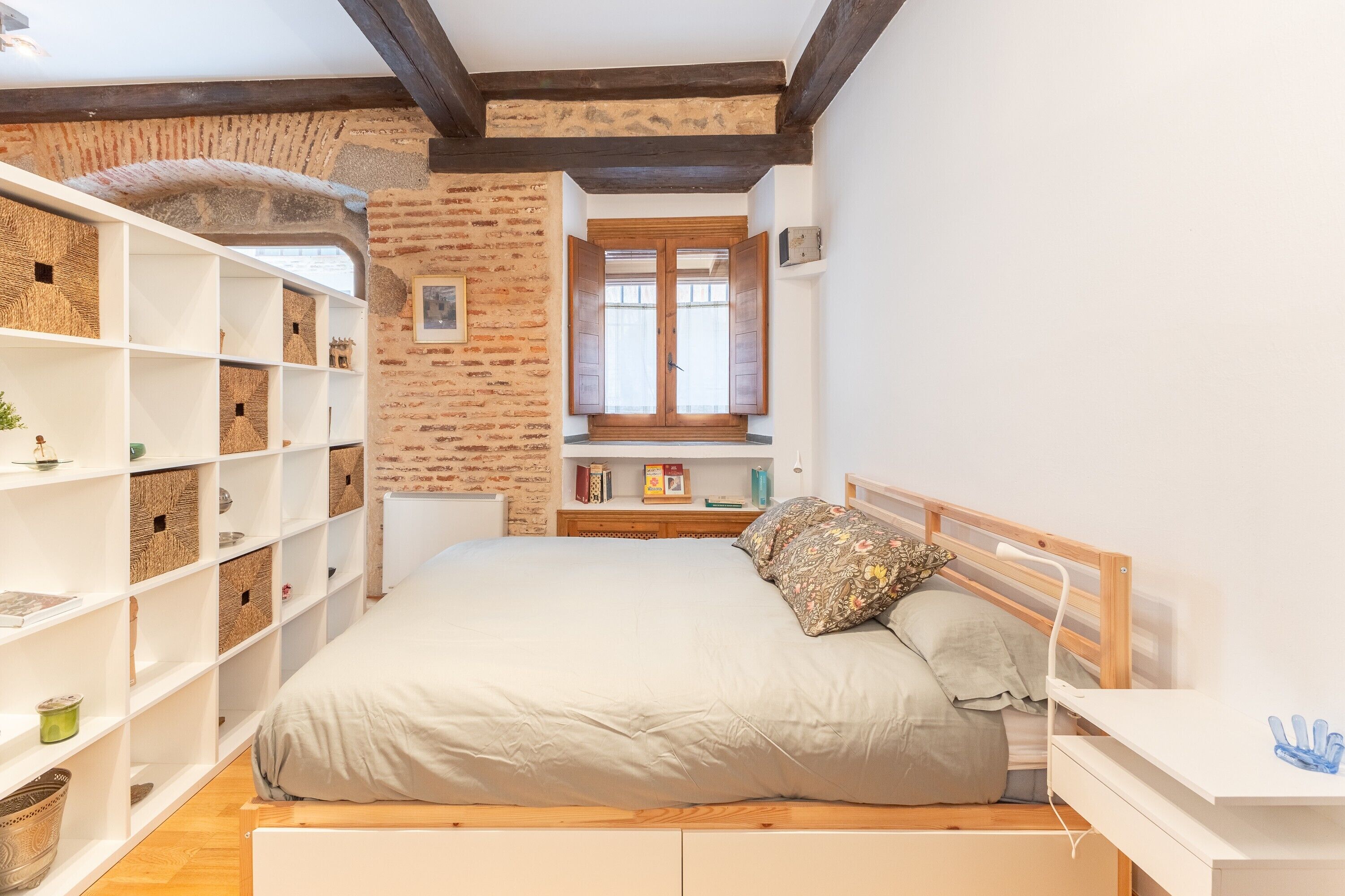 Een slaapkamer met een bed, een houten hoofdbord, een witte nachtkastje en een raam met houten luiken.