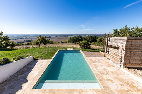 Una piscina rectangular con agua cristalina, rodeada por una solana empavedada, con un paisaje rural de campos y árboles de fondo.