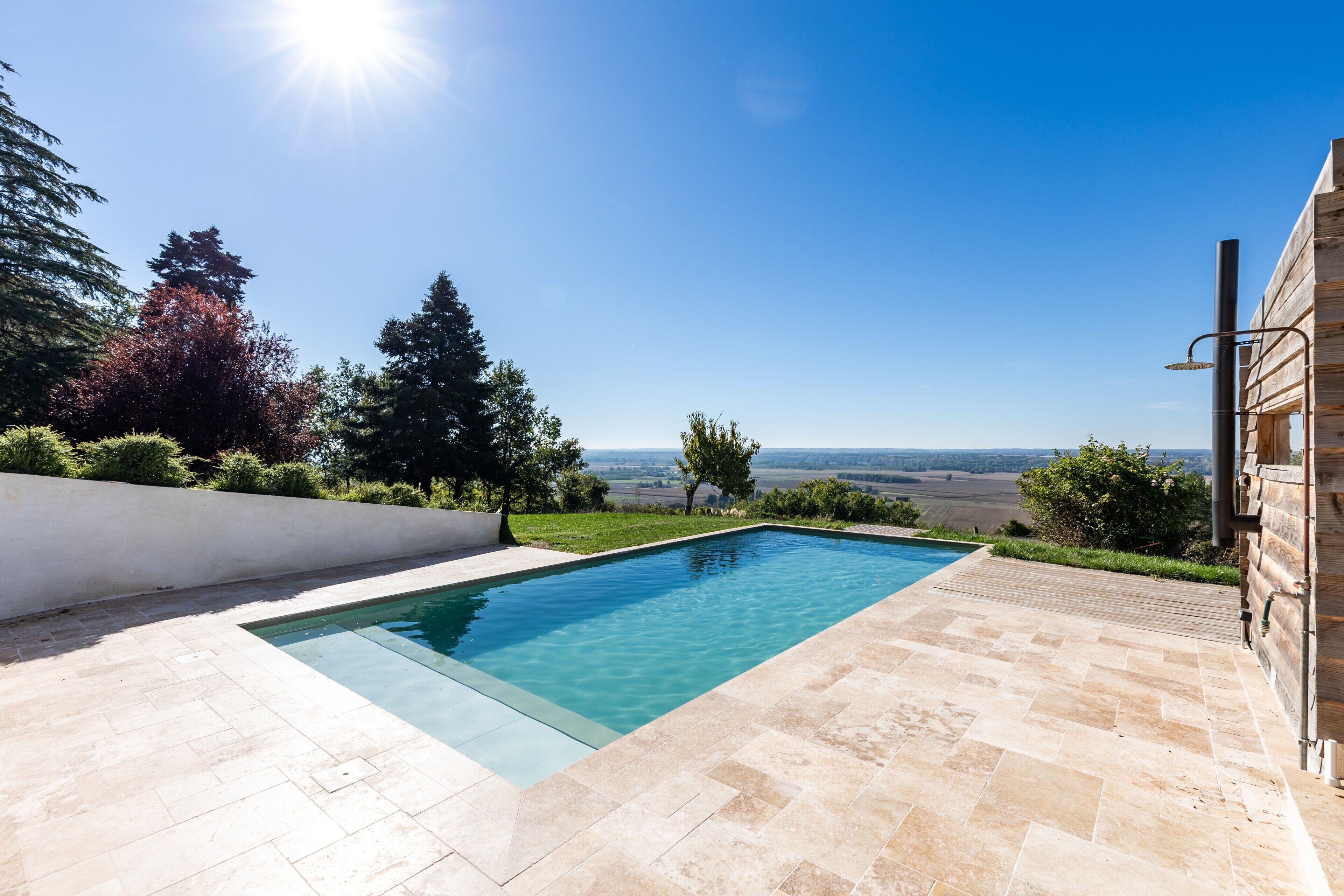 A rectangular swimming pool with clear water, surrounded by beige tiles, set against a backdrop of trees and a clear sky.
