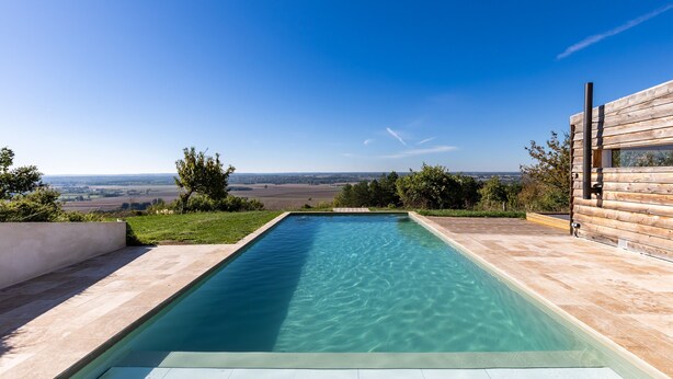 A rectangular swimming pool with clear water, surrounded by a tiled deck and a wooden building with a window.