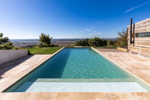 Una piscina rectangular con agua cristalina, rodeada por una solana con baldosas y un edificio de madera con una ventana.
