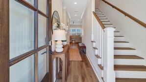 A hallway with a wooden staircase, a mirror, a lamp, and a rug leading to a living area.