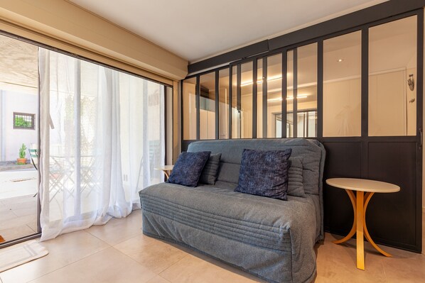 A room with a grey sofa, a small round table, and a sliding glass door leading to an outdoor area.