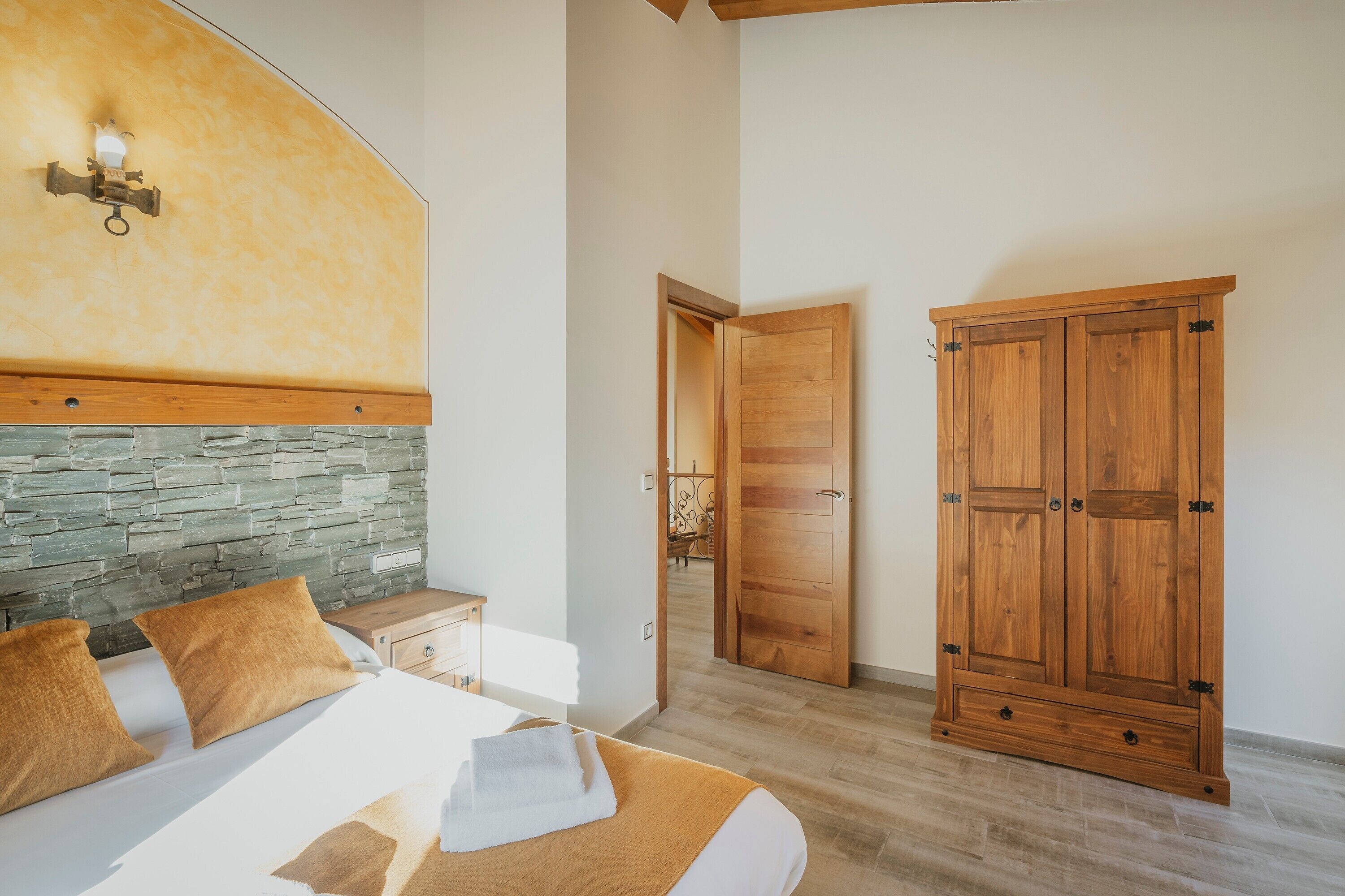 A bedroom with a stone headboard, a wooden wardrobe, and a bed with a white comforter and brown pillows.
