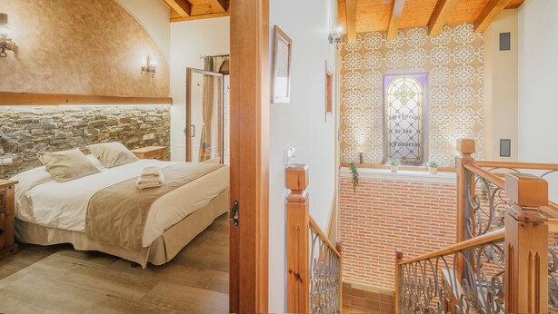 A bedroom with a stone wall, wooden ceiling beams, a bed, and a staircase leading to a lower level.
