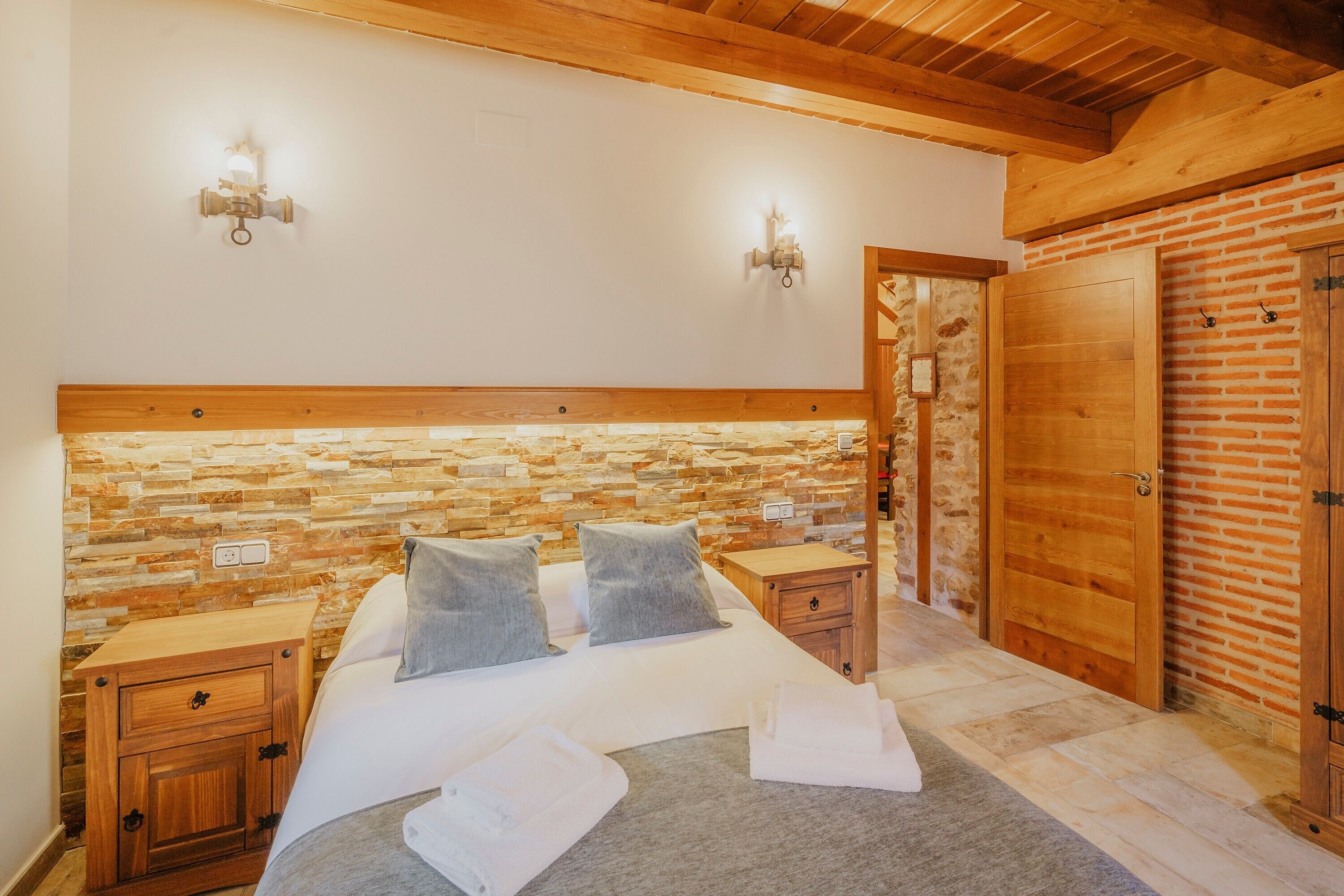 Un dormitorio con pared de piedra, cabecero de madera, dos mesitas de noche, una cama con almohadas y un armario de madera.