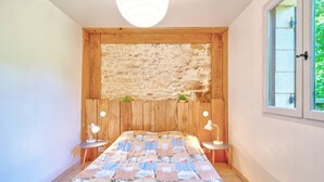 A bedroom with a stone wall, wooden headboard, two bedside tables with lamps, a bed with a patterned bedspread, and a window with a view. - Charming Holiday Home Quiétude in Meyrals (Meyrals)