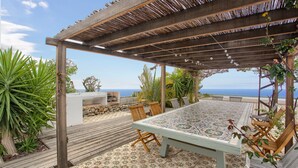 Een overdekt terras met een tafel en stoelen, omgeven door planten en uitzicht op de zee.