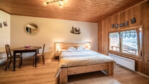 Une pièce aux murs lambrissés, comprenant un lit, un miroir rond, une table à manger avec des chaises et une fenêtre avec des rideaux.
