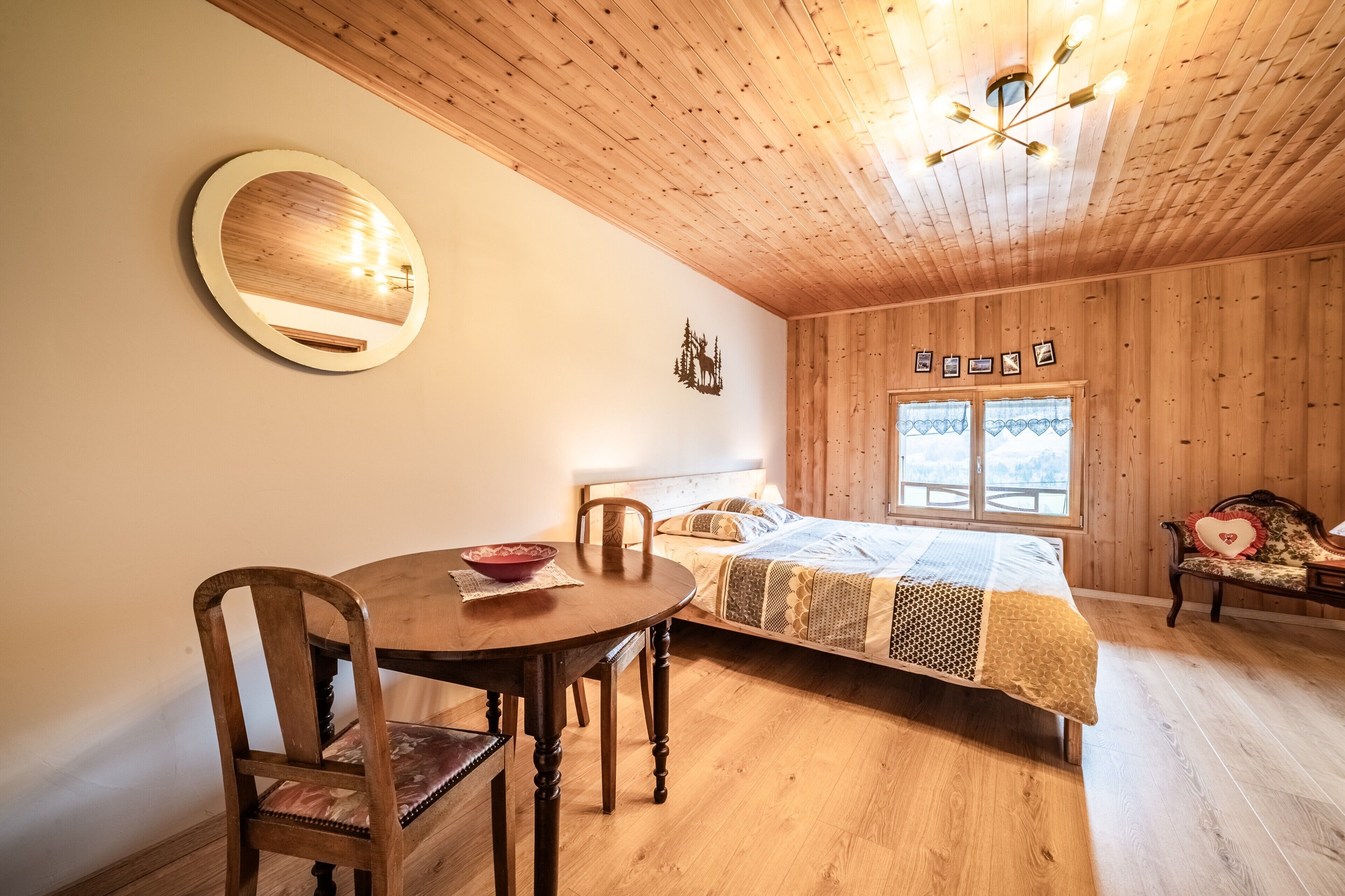Una habitación con paneles de madera, una cama, una mesa de comedor con sillas, un espejo redondo y una araña.