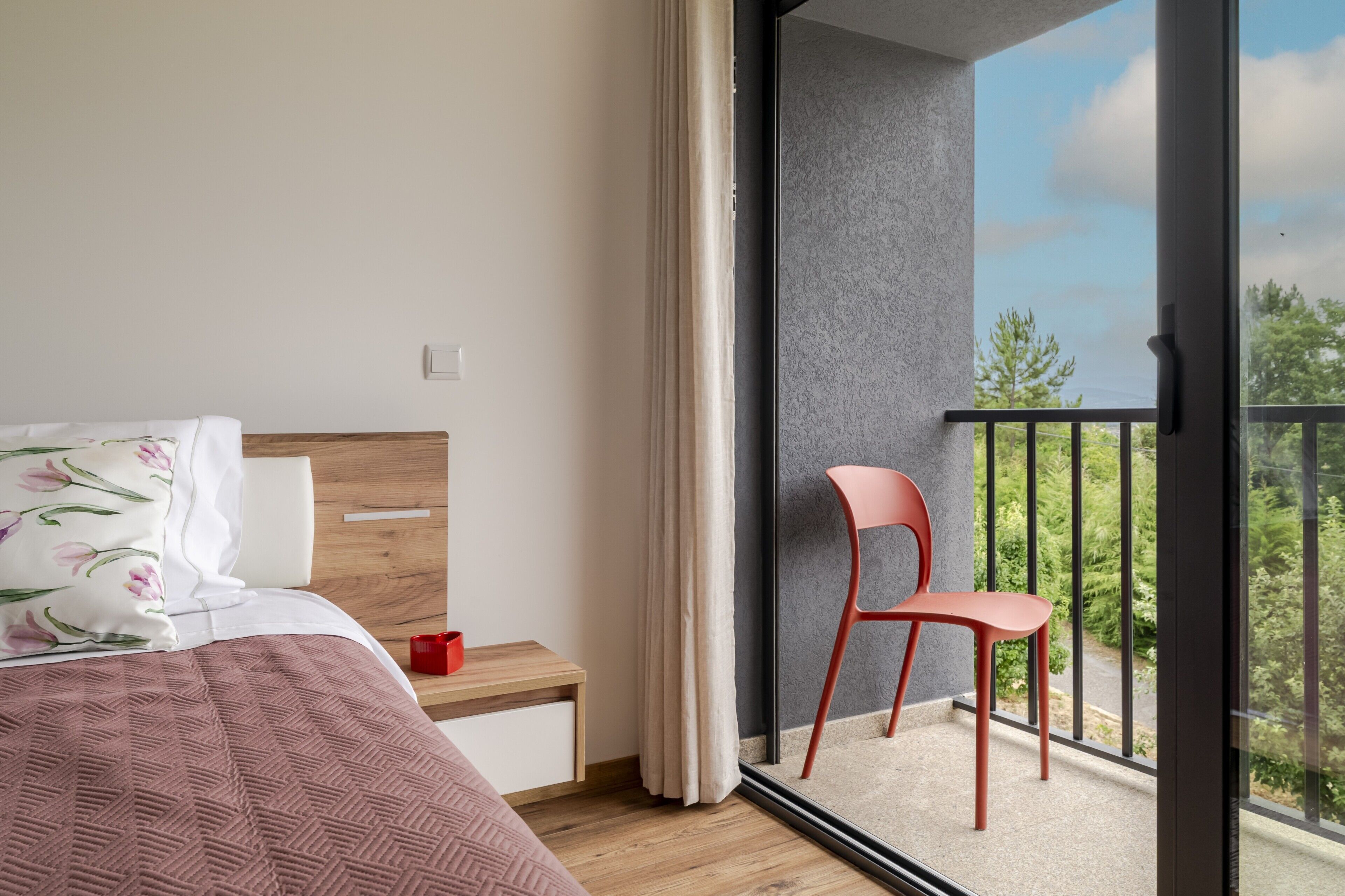 Ein Schlafzimmer mit Bett, Nachttisch, Sessel und einem Balkon mit Blick auf Bäume.