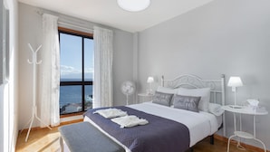 Un dormitorio con cama, mesitas de noche, un banco, vista al mar y una ventana con cortinas.