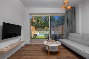 A modern living room with a flat-screen TV, a sofa, a dining table, and a view of a backyard with a pool.