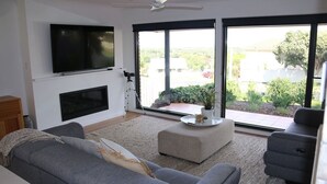 Ein modernes Wohnzimmer mit einem grauen Sofa, einem Couchtisch und einem an der Wand montierten Flachbildfernseher. Eine Schiebetür aus Glas führt zu einem Balkon mit Blick auf Grünflächen und Gebäude.