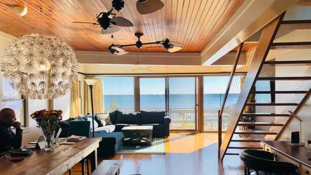 A spacious living room with a wooden ceiling, a large chandelier, and a view of the ocean through floor-to-ceiling windows. - 3 bedroom, waterfront cottage on the shores of the Chesapeake Bay. (North Beach)