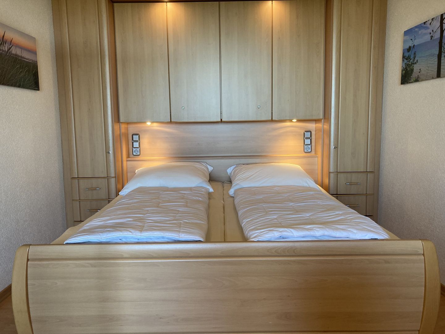 Un lit double avec une tête de lit en bois et deux tables de chevet, une armoire en bois et deux interrupteurs d’éclairage fixés au mur.