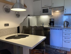 A compact kitchen with a white countertop, a microwave, a coffee maker, and a sink. There are two chairs and a table with a placemat.