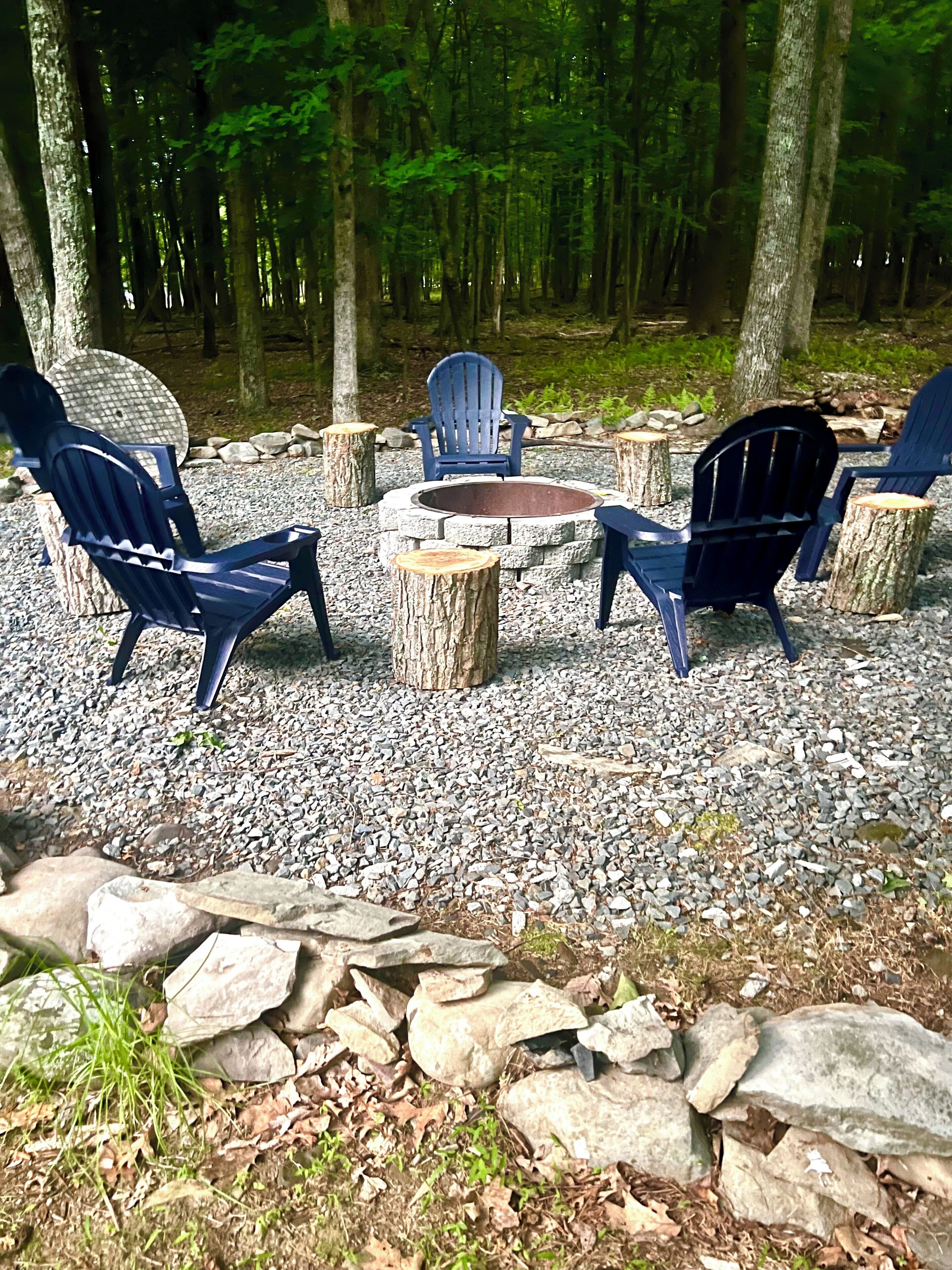 A forest setting with a fire pit and four Adirondack chairs arranged around it.