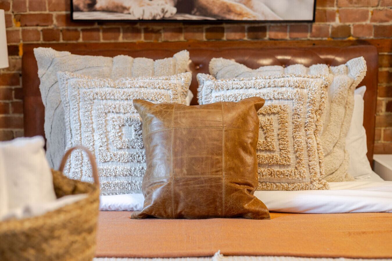 Una cama con un cojín de cuero marrón, almohadas blancas con textura y un cabecero de madera frente a una pared de ladrillo.