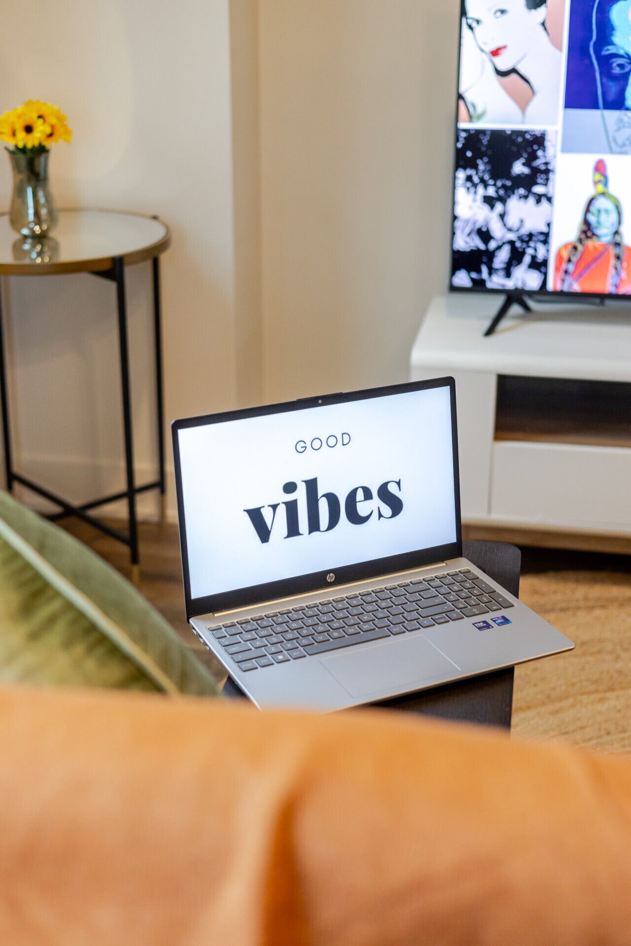 A laptop on a table displaying 'GOOD vibes' with a TV in the background.