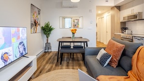Un soggiorno moderno con televisione a schermo piatto, un tavolo da pranzo con sedie, un divano grigio con cuscini, una pianta in vaso e un angolo cucina con elettrodomestici.
