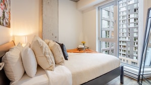 Camera da letto con un letto, cuscini, un comodino con una lampada, una finestra vista edifici e una parete in cemento.