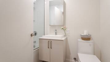 Une salle de bain moderne avec une douche en verre, un lavabo blanc avec un miroir et des toilettes, ainsi qu’un panier contenant des articles de toilette.