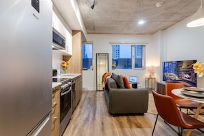 A modern kitchen with a refrigerator, oven, and microwave. There is a dining area with a table and chairs, and a sofa with a TV.