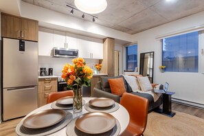 A modern kitchen with a refrigerator, microwave, and coffee maker. There is a dining area with a table set for four, a sofa, and a laptop on a side table. - Cute 1BDR Apt; Ste-Catherine - MTL Live Spot (Montréal)