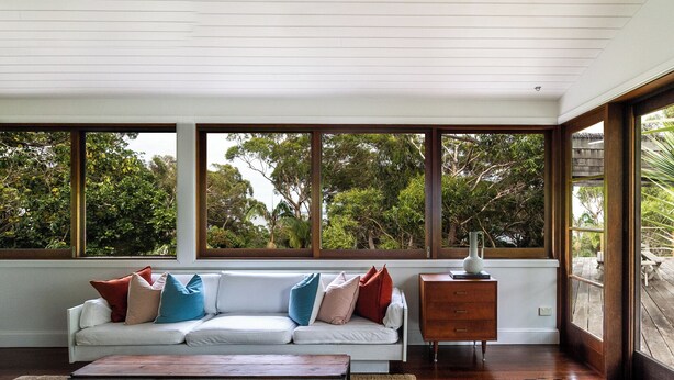 A modern living room with a white sofa, wooden floor, and large windows offering a view of trees.