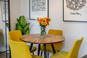 Una mesa redonda de madera con un jarrón de flores, rodeada de sillas amarillas, en una habitación con cuadros en las paredes.