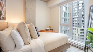 Une chambre dotée d’une grande fenêtre, d’un lit avec une literie blanche, d’un mur en béton servant d’élément décoratif, et offrant une vue sur un immeuble.
