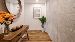 Un couloir avec un banc en bois, un miroir rond, un vase avec des fleurs, un code QR encadré et une plante en pot.