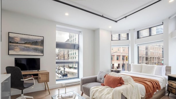 A modern bedroom with a bed, a desk, a chair, and a large window with a city view.