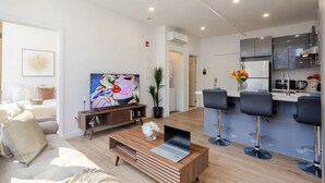 Una sala de estar moderna con un sofá, una mesa de centro, un televisor y una zona de cocina con taburetes de bar.