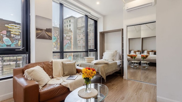 Una sala moderna con un sofá de cuero marrón, una mesa de centro de vidrio con un florero, una cama con ropa de cama blanca y ventanas amplias que ofrecen vistas a la ciudad.