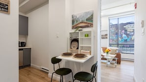 Una cocina compacta con electrodomésticos modernos, un comedor con dos sillas y vistas a una pizzería a través de un ventanal grande.