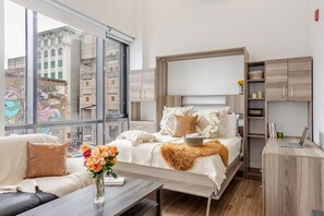 A modern bedroom with a large window, a bed with multiple pillows, a bedside table with a vase of flowers, and built-in wooden storage.
