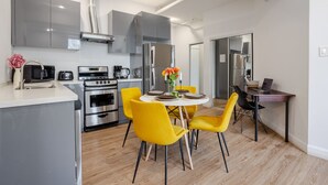 Une cuisine moderne équipée d’appareils électroménagers en acier inoxydable, un coin repas avec des chaises jaunes et un petit espace de travail avec un ordinateur portable.