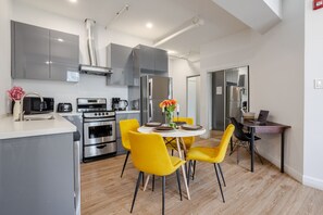A modern kitchen with stainless steel appliances, a dining area with yellow chairs, and a small workspace with a laptop.