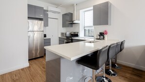 A modern kitchen with grey cabinets, a stainless steel refrigerator, a microwave, and a stove. There is a bar area with black barstools.