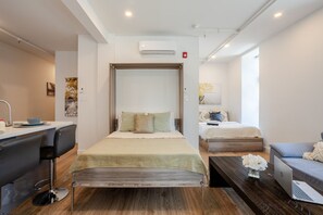 A modern hotel room with a bed, a desk, a chair, a sofa, and an air conditioning unit.